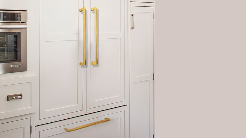 White kitchen cabinets with gold handles, including two vertical handles on the central doors and a horizontal handle on a bottom drawer; part of an oven is visible on the left.