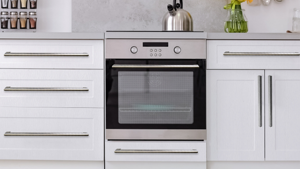Modern kitchen with a stainless steel oven built into white cabinets, a kettle on the countertop above, jars of spices on the left, and a green vase with yellow flowers on the right.