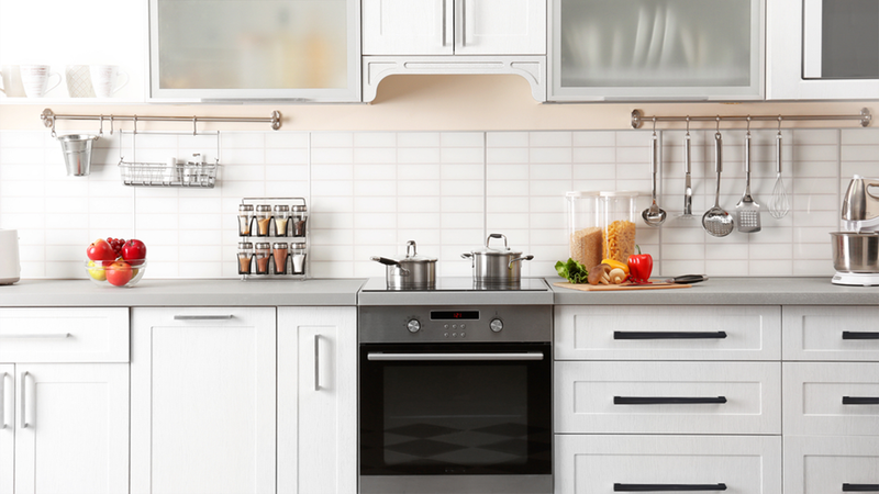 Modern white kitchen with a built-in oven, stove, and countertop holding pots, jars of pasta and lentils, apples, and kitchen utensils hanging on the wall above white cabinets.
