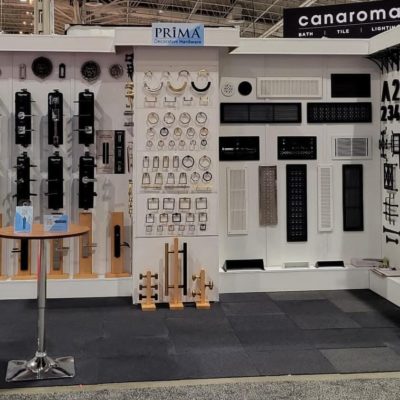 Display booth featuring various hardware items such as door handles, grilles, and vents arranged on a white wall, with a small round table in front and signage for PRiMA and canaroma above.
