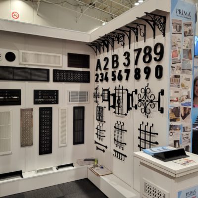 Display wall at a trade show booth featuring black metal house numbers, decorative grilles, vents, and ornate brackets. Brochures and a display stand for PRIMA Home Hardware are visible on the right.