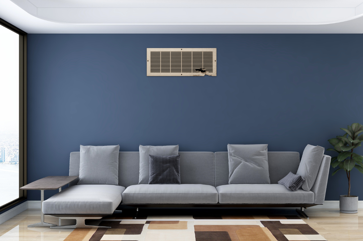 A modern living room with a gray sectional sofa, blue accent wall, and large window. A broken air vent is mounted high on the wall. There’s a potted plant and a rug with geometric patterns on the floor.