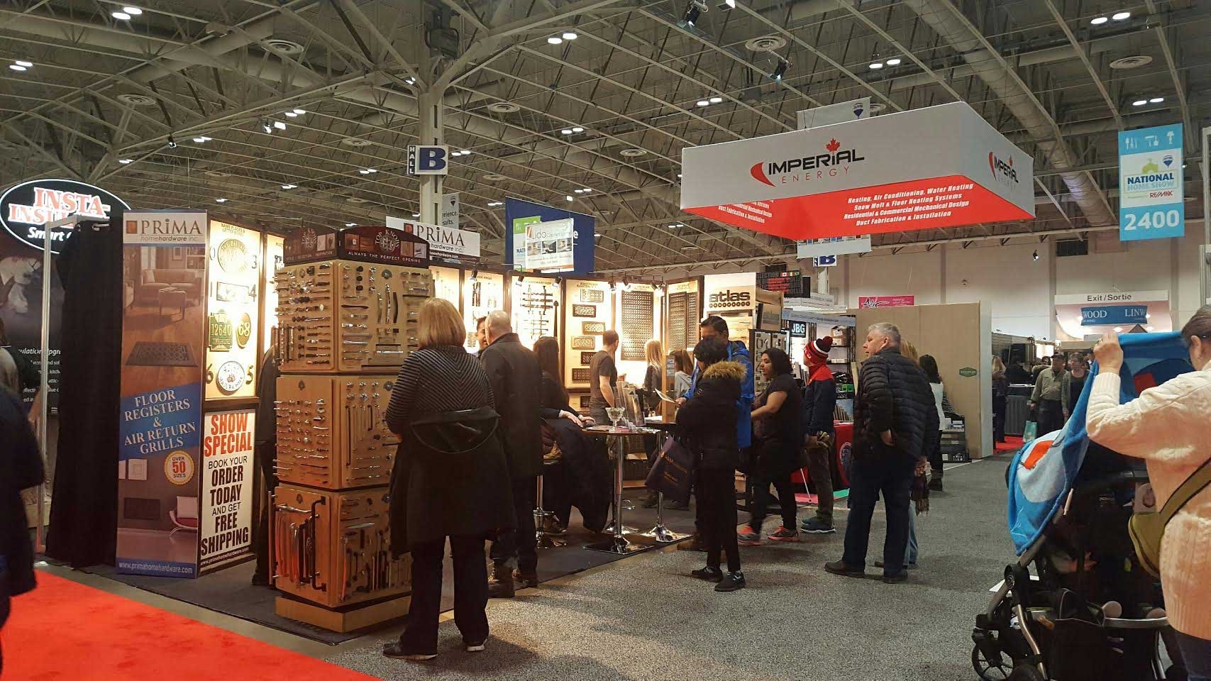 A busy trade show floor with various booths, signs, and displays. People are walking, browsing, and interacting with exhibitors under high ceilings with visible lights and banners.