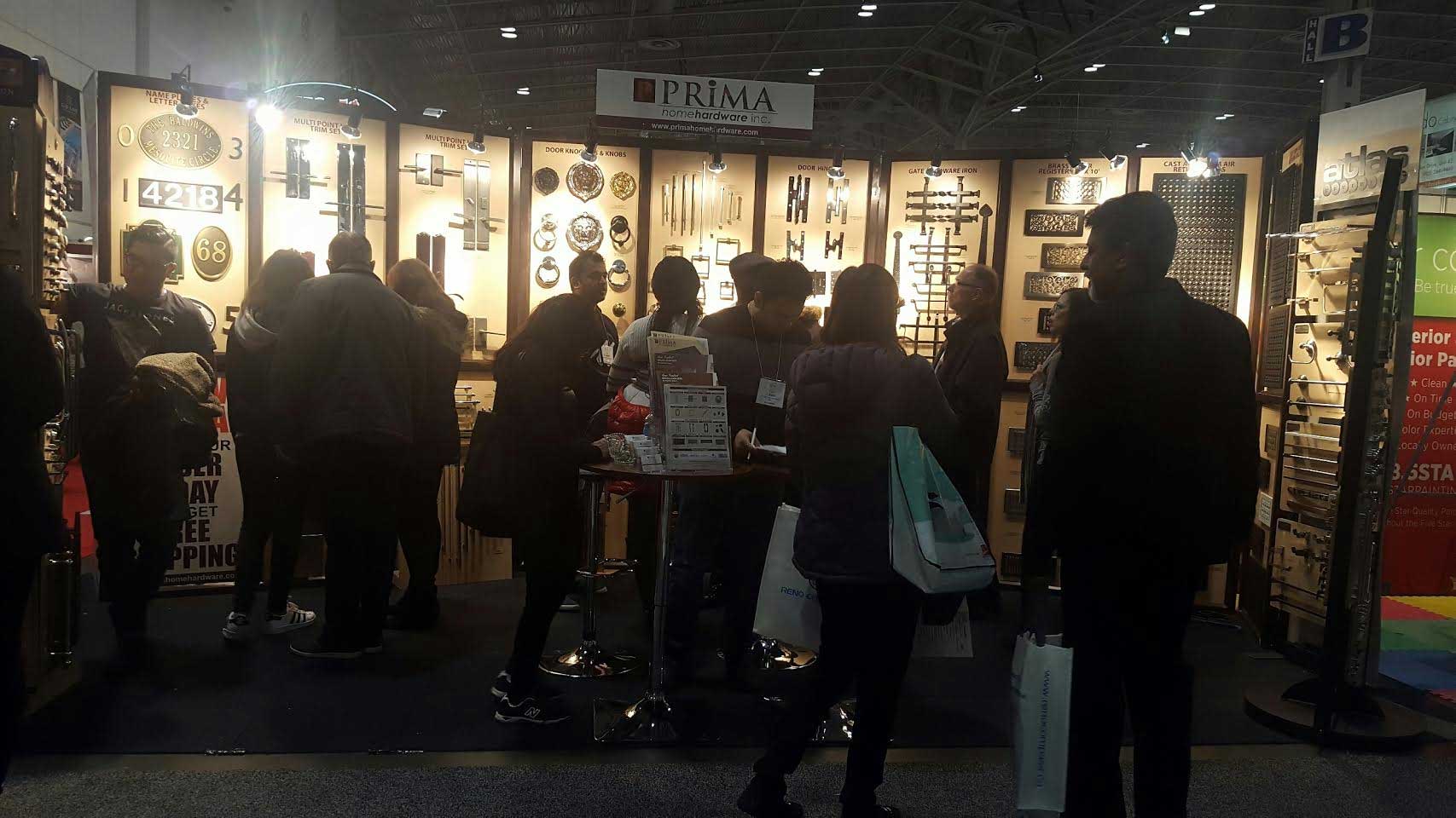 People gather at a trade show booth displaying various metallic hardware items, such as handles and locks, on wooden panels. Some attendees are engaging with exhibitors, holding shopping bags and flyers. The booth is labeled PRIMA.