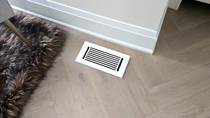A rectangular white floor vent with black slats is installed in light wood flooring near a white baseboard and a brown furry rug. The corner of a chair leg is visible on the left side.
