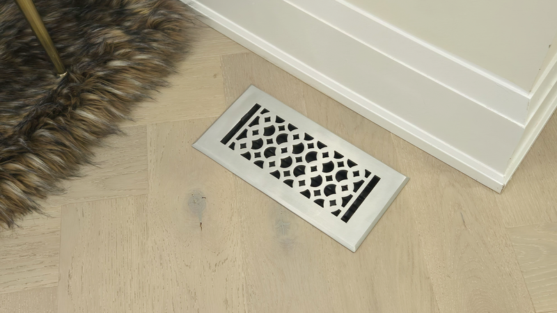 A decorative metal floor vent cover with a geometric pattern is set in light-colored, herringbone-patterned wood flooring near a white baseboard and a fuzzy brown rug.