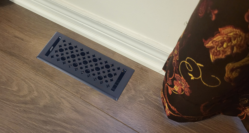 A decorative black floor vent cover is installed on brown wooden flooring next to a white baseboard, with a patterned brown curtain partially visible on the right.