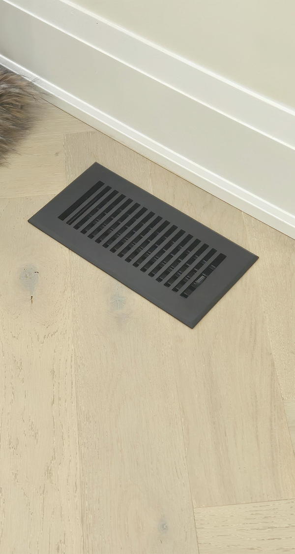 A black rectangular floor vent is set into light-colored wooden flooring near a white baseboard. A small patch of fur or a pets tail is visible in the top left corner.