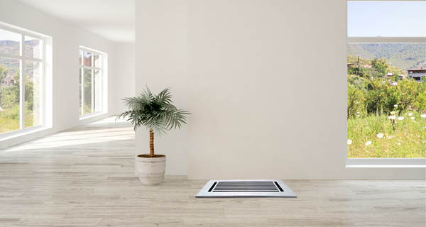A small potted palm sits beside a wall vent in a bright room with large windows overlooking a green, flower-filled field and distant hills. The floor is light wood, and the walls are white.