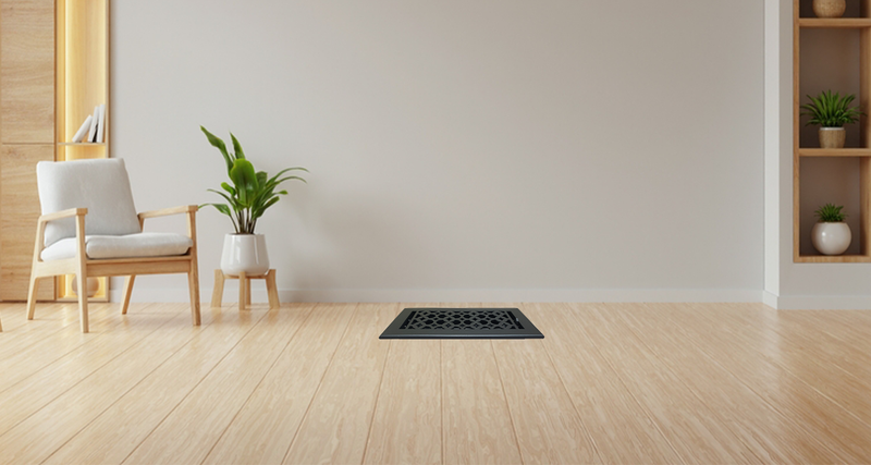 A minimalist living room with light wood flooring, a white chair with a cushion, a potted plant, built-in wooden shelves, and a decorative black vent cover on the floor against a plain light-colored wall.
