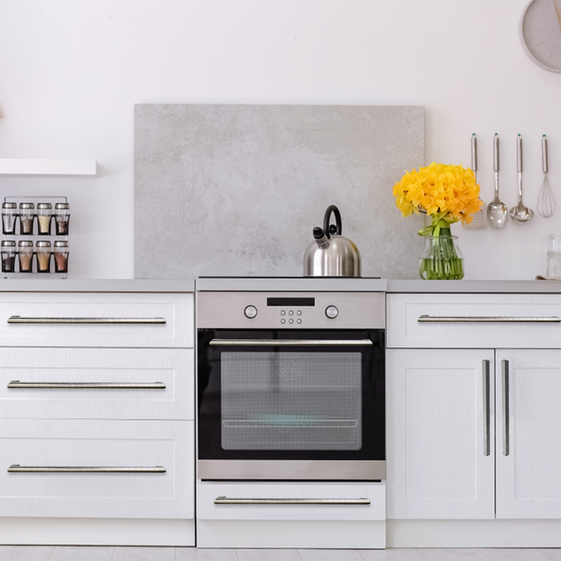 Modern white kitchen with stainless steel oven, a kettle on the stovetop, a jar of yellow flowers on the counter, spice jars on a shelf, and kitchen utensils hanging on the wall.