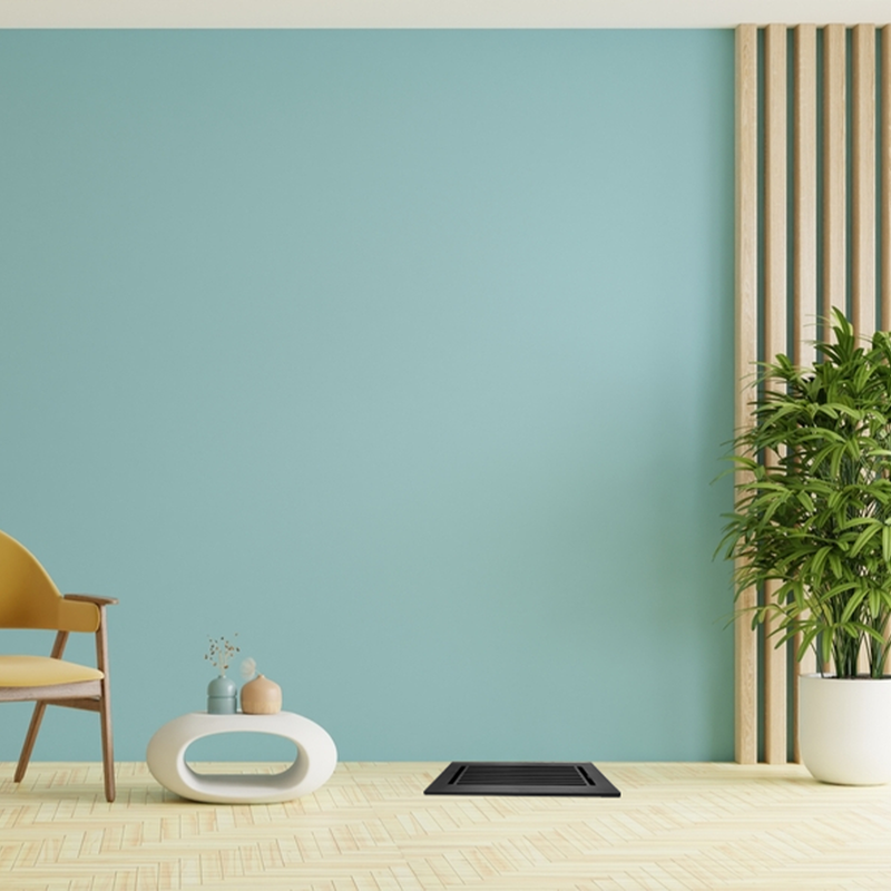 A modern living room with a light blue wall, wooden chair with a yellow cushion, white oval coffee table, large green potted plant, and wood slat decor accent. Light wood flooring and a floor vent are also visible.