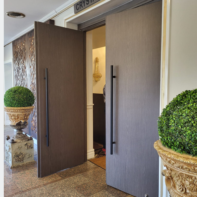 Two large wooden doors with long black handles are open, revealing part of an interior hallway with a decorative wall sconce. Ornate planters with trimmed green topiary plants stand on either side of the entrance.