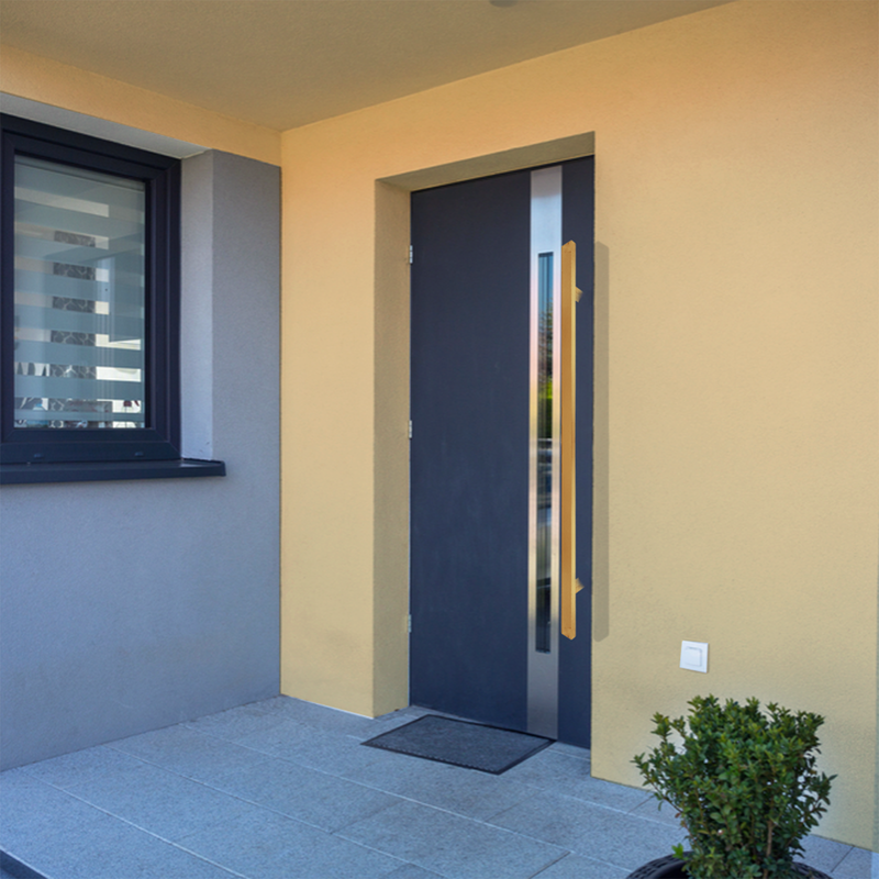 Modern front door with a vertical handle, set in a yellow and gray exterior wall, next to a window with horizontal blinds. There is a small doormat and a potted plant by the entrance.