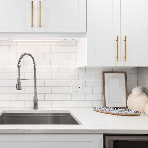 A modern white kitchen with a stainless steel sink, a pull-down faucet, white cabinets with gold handles, a white subway tile backsplash, and decorative items on the counter.