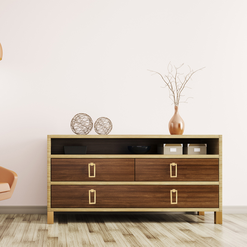 A modern dresser with four drawers sits against a light wall. On top are two decorative globes, a bowl, two storage boxes, and a tall vase holding bare branches. Part of an orange chair is visible on the left.