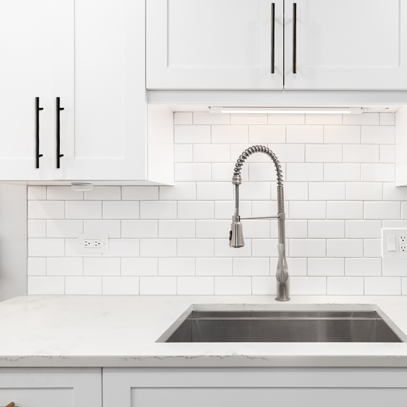 Modern kitchen with white cabinets, subway tile backsplash, marble countertop, and a stainless steel sink featuring a high-arc pull-down faucet. Black cabinet handles provide a contrasting accent.