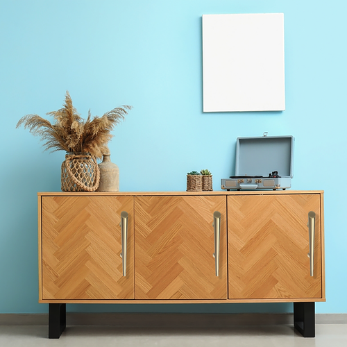A light wooden cabinet with chevron pattern stands against a blue wall. On top are dried flowers in a vase, small potted plants, and an open blue record player. A blank white square frame hangs above.