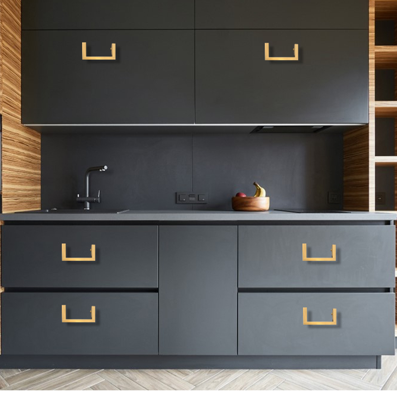 A modern kitchen with matte black cabinets, gold handles, a sink, and a bowl of fruit on the counter. The backsplash and side walls feature wood paneling, and the floor has a herringbone pattern.