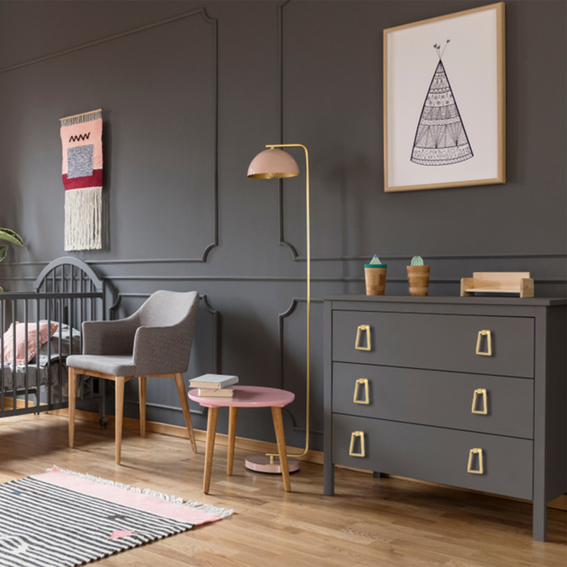 A modern nursery features a gray crib, armchair, and dresser with gold handles. A pink and gray rug, wall art, and woven tapestry add decorative touches. A pink lamp and wooden floor complete the cozy space.