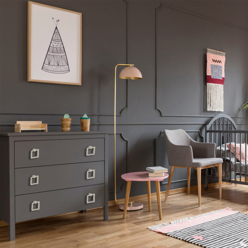 A modern nursery with dark gray walls, a framed tipi print, a gray dresser, a floor lamp with a pink shade, a gray armchair, a small pink table, a crib, and a woven wall hanging. Wooden floor with a striped rug.