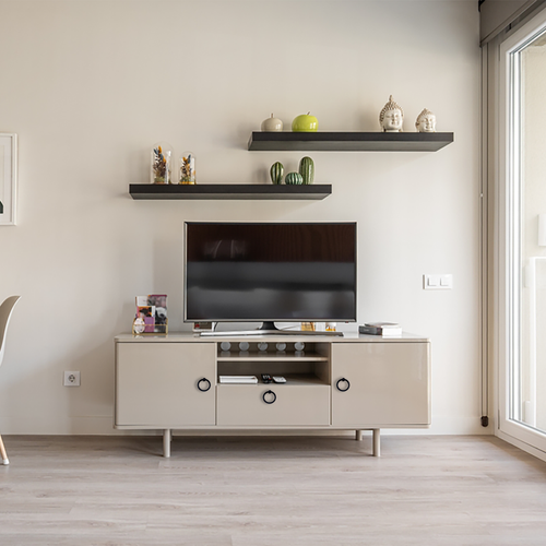 A modern living room setup with a flat-screen TV on a beige cabinet, two dark floating shelves above holding decorative items, and light wood flooring. A large window lets in natural light from the right.