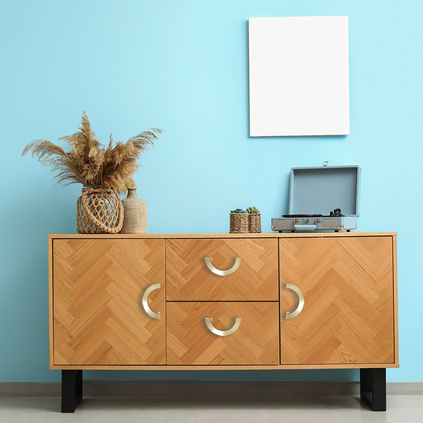 A wooden sideboard with decorative handles stands against a light blue wall. On top are a vase with dried grasses, small potted plants, and an open blue record player. A blank white frame hangs above.