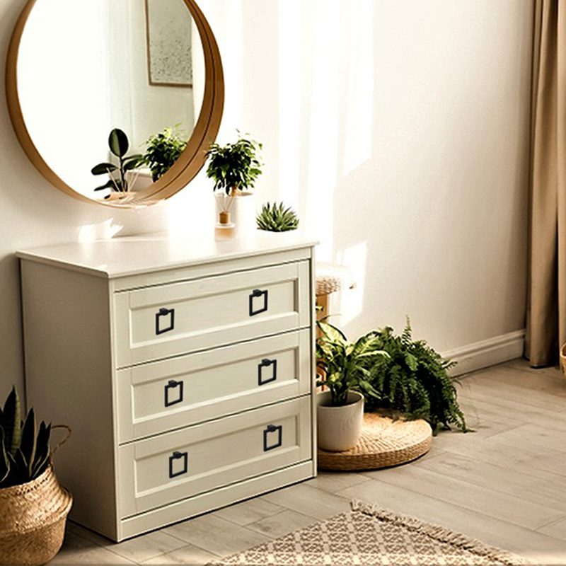 A white dresser with three drawers sits below a round mirror. Several potted plants and a small candle are on and around the dresser, with neutral decor and sunlight streaming into the cozy, minimalist room.