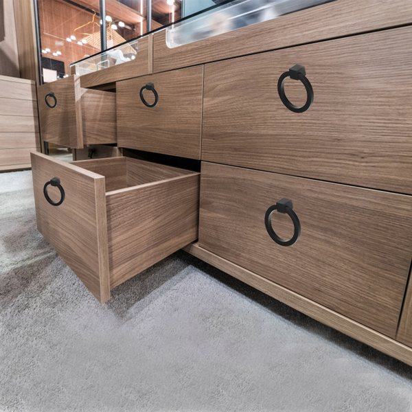 A close-up view of a wooden dresser with black circular handles, featuring one drawer partially open, situated on a carpeted floor.