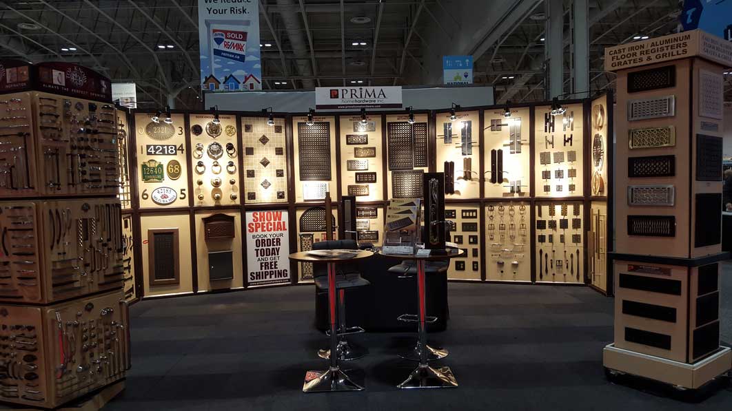 A trade show booth displays various cabinet hardware, decorative panels, metal grilles, and accessories on illuminated walls. Two high tables with stools are positioned at the front for visitors.