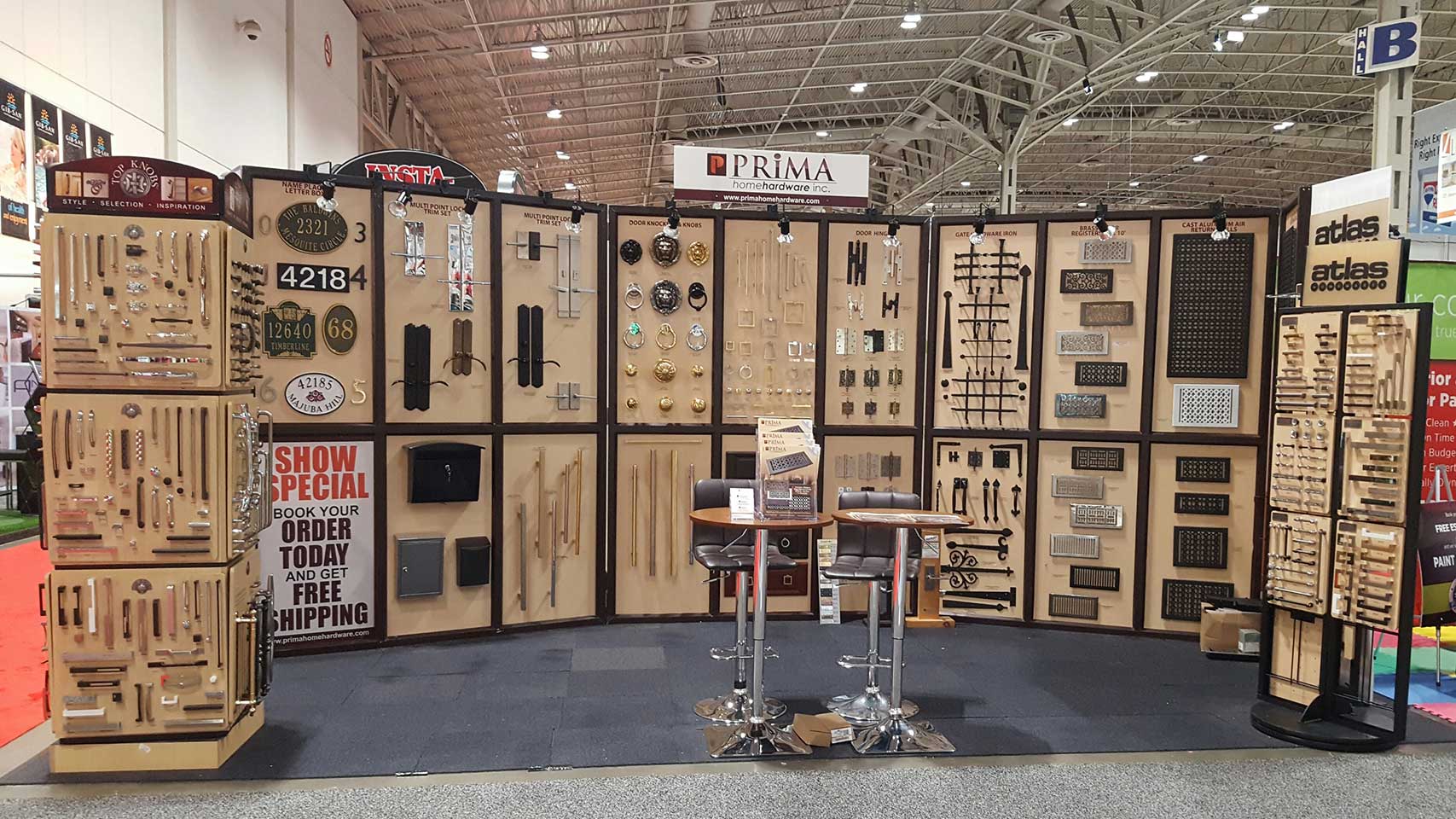 Trade show booth displaying a wide variety of cabinet hardware, including handles, knobs, and pulls, neatly arranged on boards. Two high stools and a small table are positioned in front of the display.