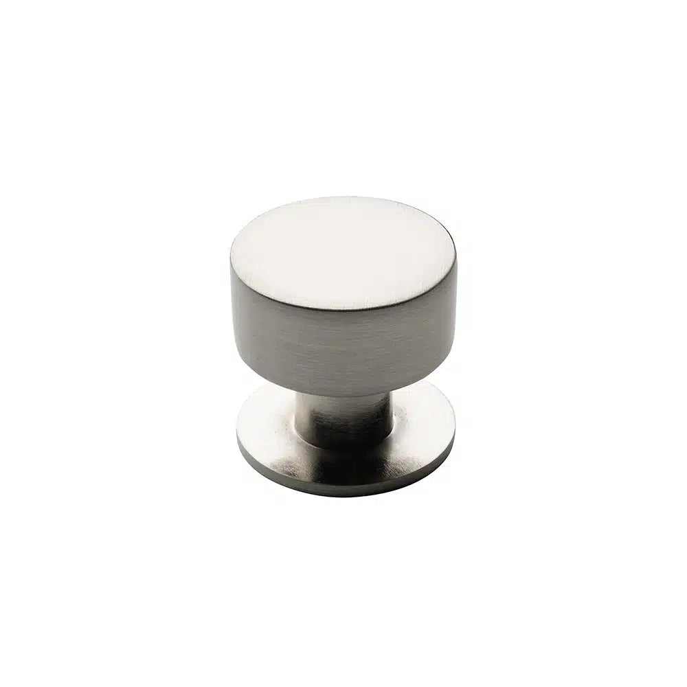 A Taymor Industries brushed nickel cabinet knob, made of durable Zinc Die-Cast, features a round flat top and a short cylindrical stem on a round base, displayed against a plain white background.