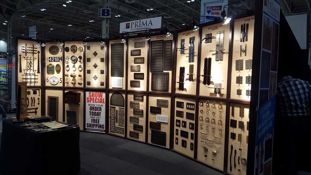 A trade show booth displays various metal hardware items such as handles, grids, house numbers, and vents, neatly arranged on illuminated panels. A sign advertises special offers and free shipping.