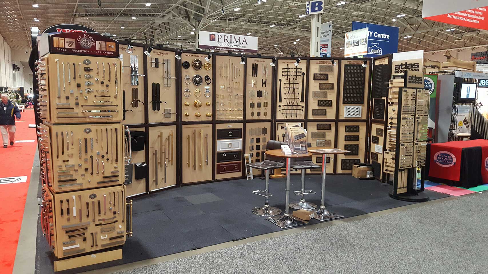 Display booth at a trade show featuring various hardware items such as handles, knobs, and locks mounted on boards, with chairs and a small table in the center of the booth. Signs and banners are visible in the background.