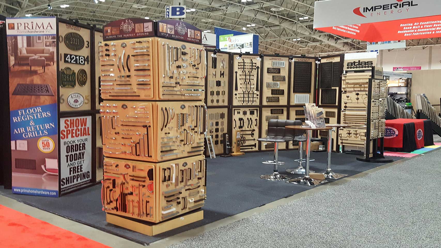 Trade show booth displaying various metal hardware pieces like handles and knobs on boards and rotating stands, with banners, sample boards, a table, and chairs on a carpeted floor in a convention hall.