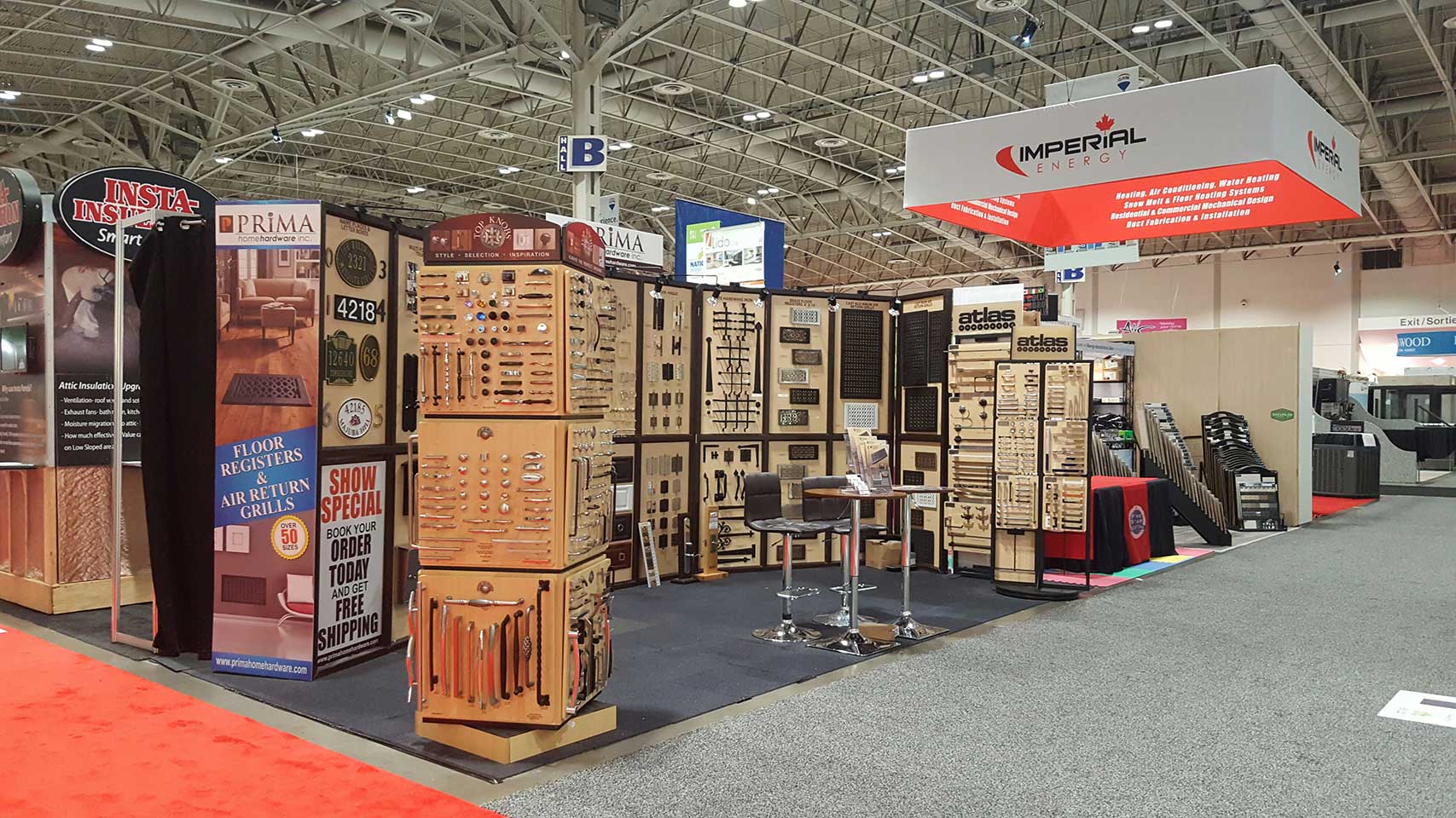 A trade show booth displays various flooring, cabinet, and hardware samples on wooden racks and panels, with signs highlighting product features and promotions. Two tall chairs and a small table are set up in the center.