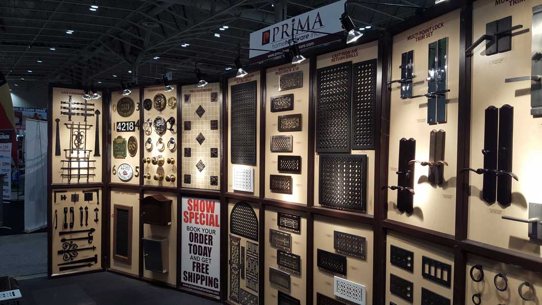 A trade show booth displays various metal architectural hardware pieces on wall panels, including grilles, vents, and decorative items. A sign advertises a show special with free shipping. The booth is well-lit and organized.