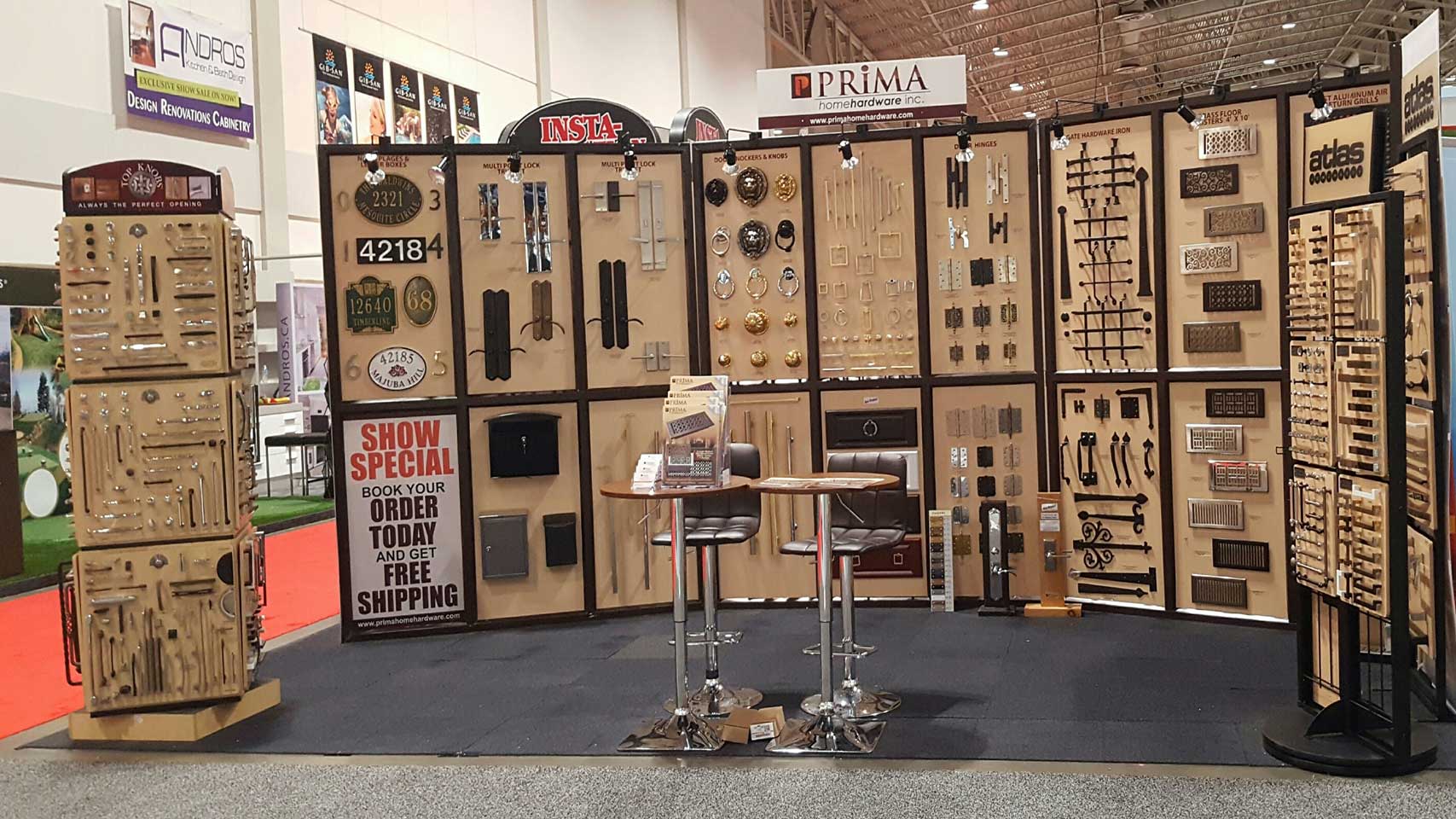 A trade show booth displays various cabinet handles, knobs, and hardware neatly arranged on tan panels. Two stools and a small table are in front of the booth, with signage promoting specials and free shipping.
