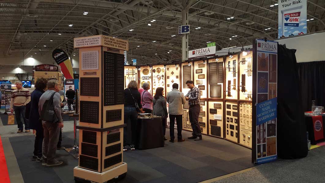 People look at decorative vent covers and grilles displayed at a well-lit trade show booth. The booth features various metal panels mounted on the walls and signs advertising custom decorative hardware.