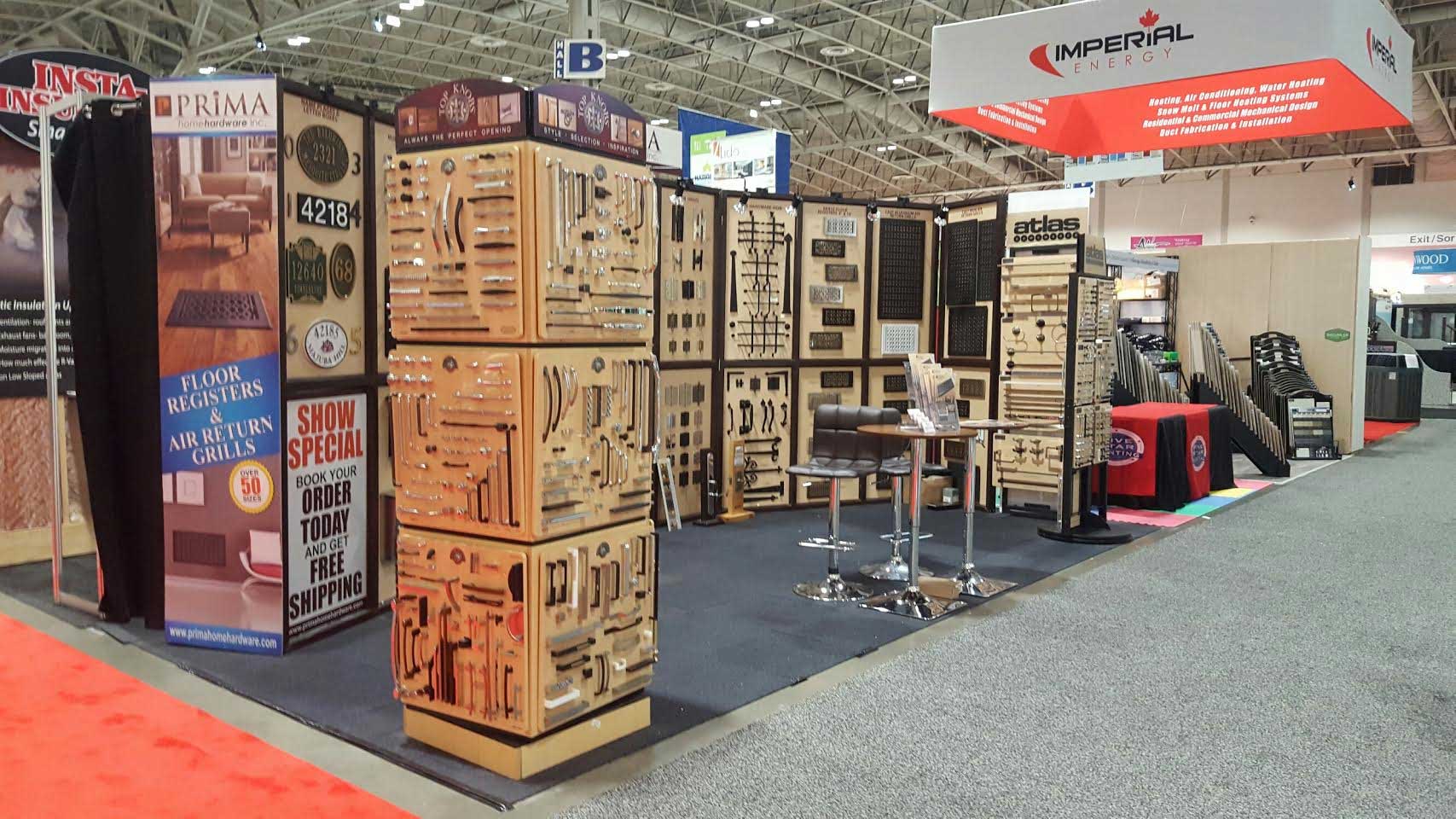 A trade show booth displays various metal hardware and decorative fixtures on wooden panels, with flyers, banners, and bar stools arranged on a carpeted floor inside a large exhibition hall.