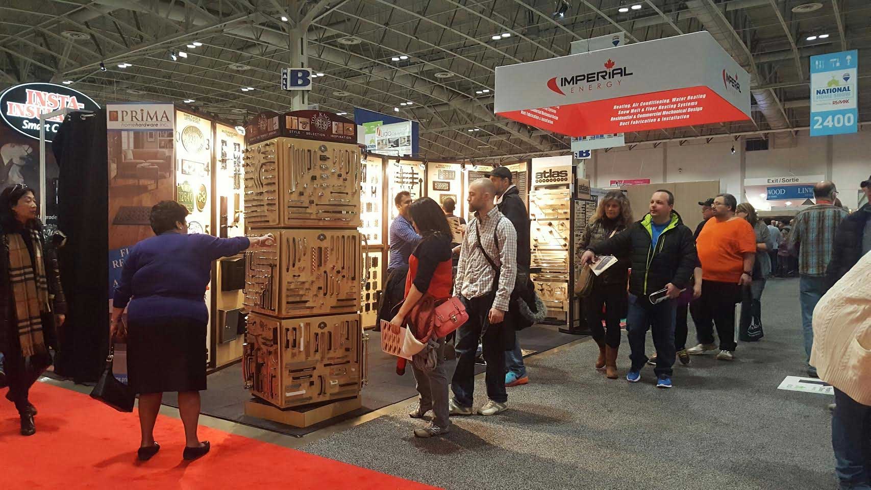 People browse a booth displaying wooden kitchen utensils at a busy indoor trade show. Several attendees walk past, while others examine products. Booth signs and bright lighting are visible overhead.