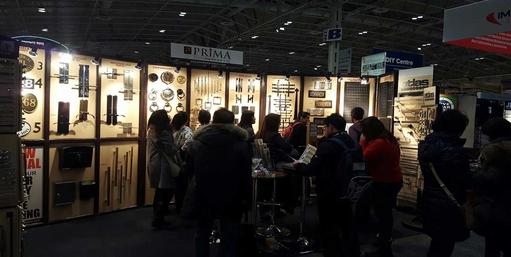 People browsing and examining hardware displays, such as door handles and locks, at a well-lit trade show booth with various products showcased on the walls.