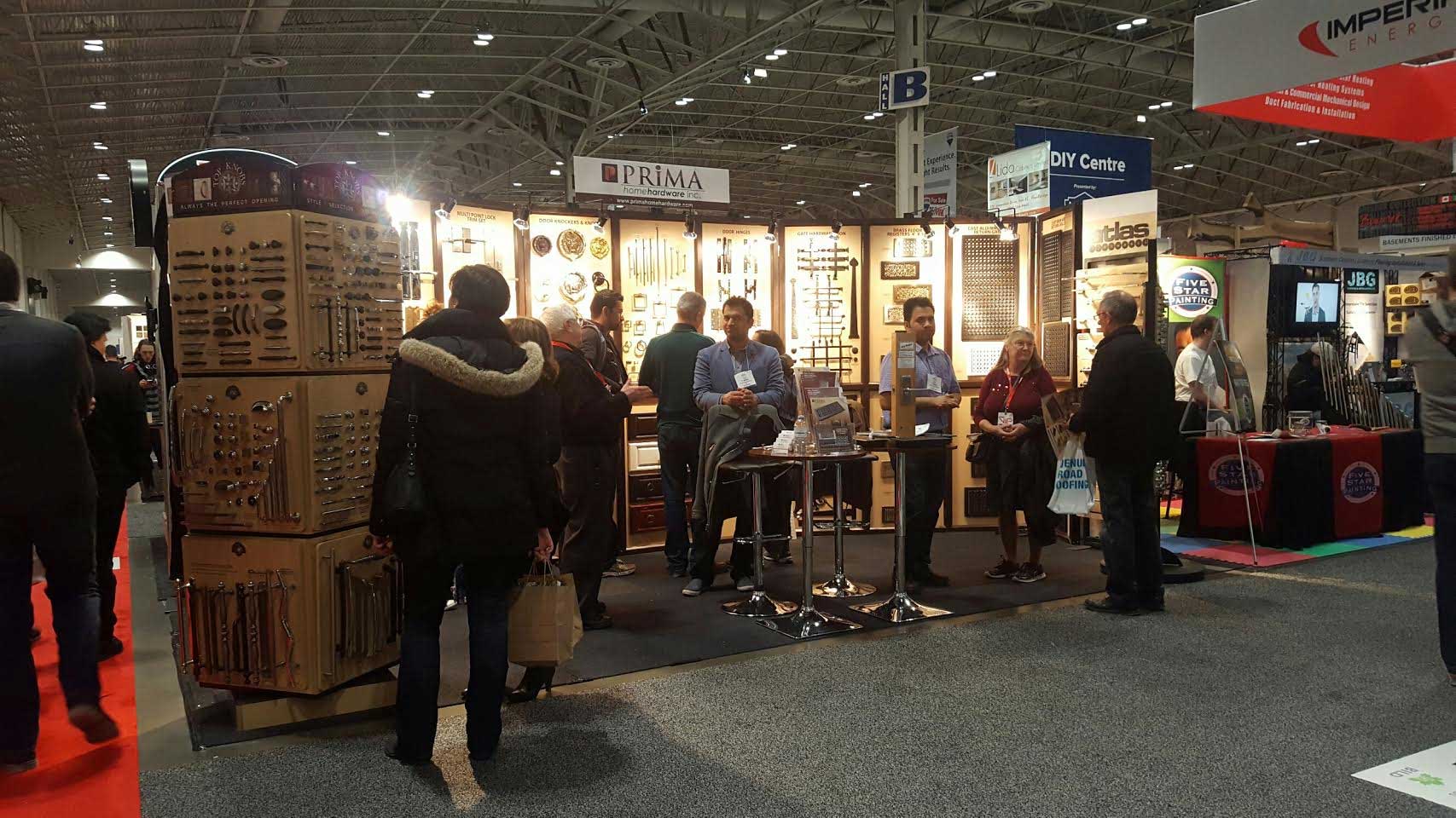 People attending a trade show or expo, gathered around a booth displaying hardware products like handles and knobs. The booth is well-lit, with informational displays and discussion among visitors and exhibitors.