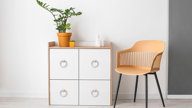 A modern, minimalistic room with a yellow chair next to a white four-drawer cabinet. A potted plant and two candles sit on top of the cabinet against a plain white wall.