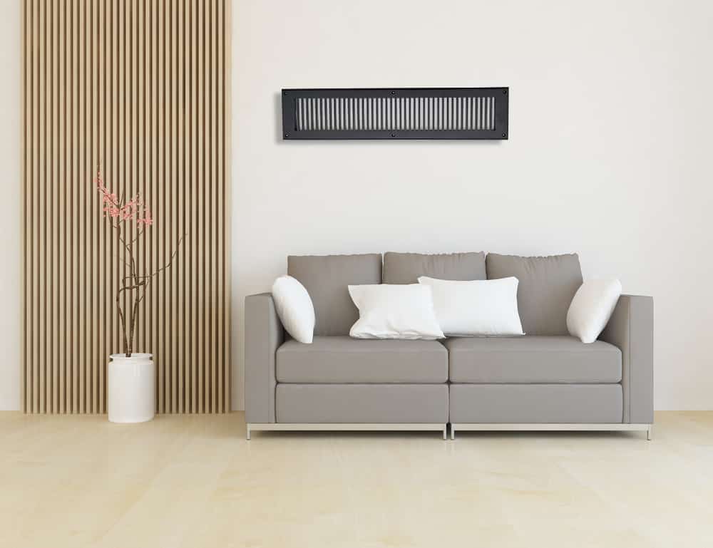 Modern living room with a light gray sofa, three white pillows, a tall vase with branches, wooden slat accent wall, and Prima Decorative Hardware cast aluminum vent covers beneath a black air conditioner on the white wall above.