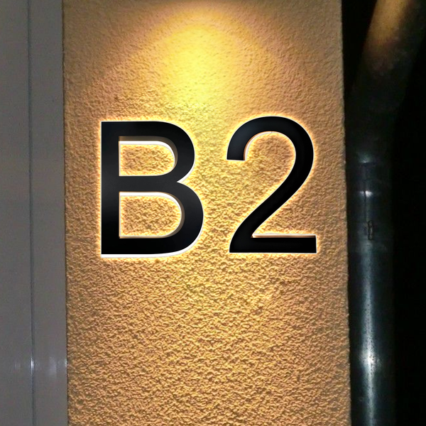 Large black letters and number B2 are illuminated on a textured beige wall, with a pipe visible on the right side. Warm lighting creates a glowing effect around the characters.