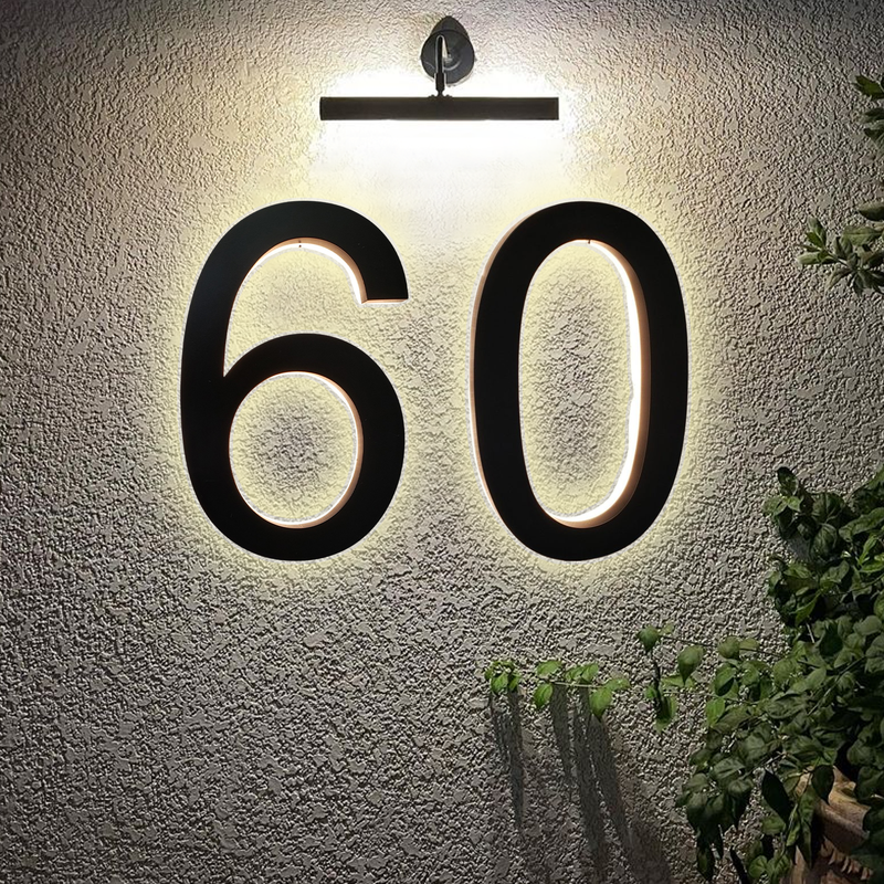 Large black numbers 60 are mounted on a textured light-colored wall, illuminated from above by a modern wall light. Green plant leaves are partially visible in the bottom right corner.