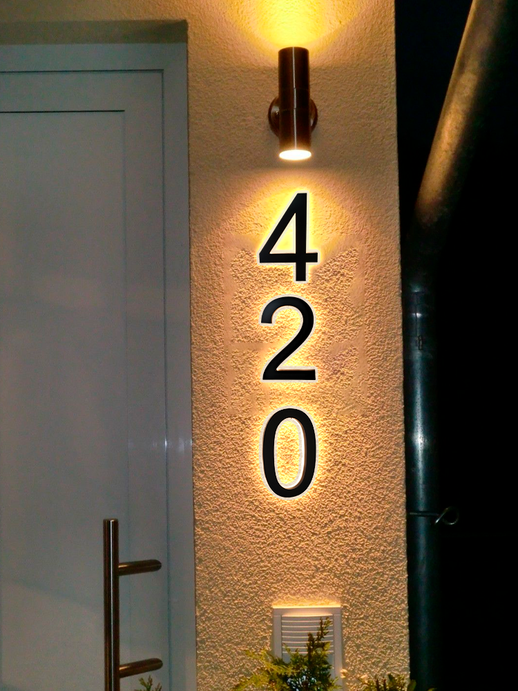 A house exterior at night with the numbers 420 mounted vertically on a lit wall next to a white door. A cylindrical light fixture above illuminates the numbers, casting shadows.