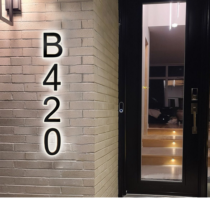 Illuminated house number B420 mounted vertically on a light brick wall next to a glass door, with warm lighting and wooden stairs visible inside the entryway.