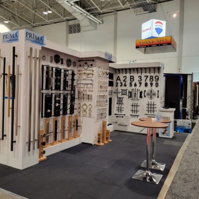 A trade show booth displays various metal hardware like rods, handles, numbers, and decorative pieces on white panels. A small round table with brochures stands in front. Signs read PRIMA and MAGNA-MAX.